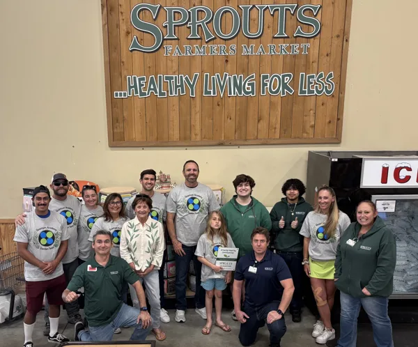 A group of people wearing Quantum Academy Garden Team shirts pose with Sprouts Farmers Market staff in front of a large wooden sign reading "SPROUTS FARMERS MARKET ...HEALTHY LIVING FOR LESS," with a young girl holding a Sprouts Foundation "Growing Healthy Communities" certificate.