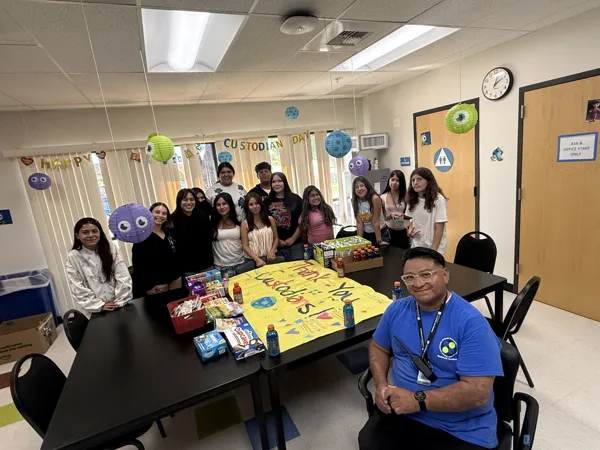 A group of students and a custodian in a blue shirt pose together in a decorated room with a yellow "Thank You Custodians!" banner and Monsters, Inc.-themed decorations for Happy Custodian Day.