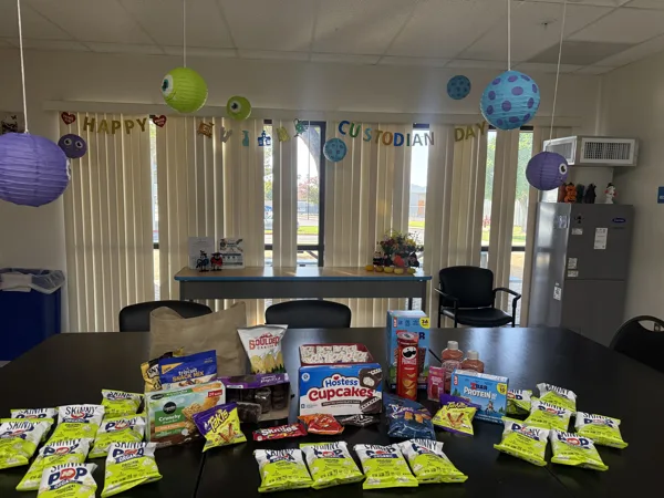 A table covered with snacks including Skinny Pop, Hostess Cupcakes, Takis, Pringles, and other treats is set up in a room decorated with Monsters Inc.-themed paper lanterns and a banner reading "HAPPY CUSTODIAN DAY."