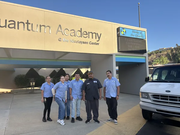 Six people in blue work shirts stand together outside Quantum Academy at the Nicolaysen Center, with a white Ford van parked nearby.