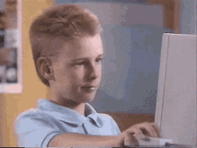 A young boy with blond hair sits at a laptop computer indoors, appearing pleased with himself.