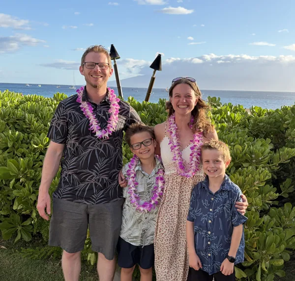 A family of four wearing flower leis poses together in front of lush greenery and the ocean on a sunny day.