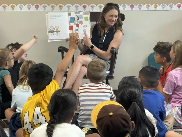 A woman sits in a rocking chair reading a picture book aloud to a group of young children seated on the floor of a classroom.