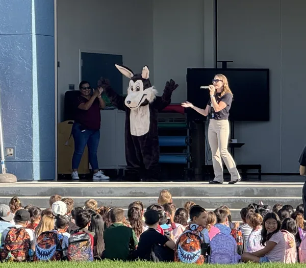 Two adults and a wolf mascot perform on an outdoor school stage in front of a large group of children seated on the grass.