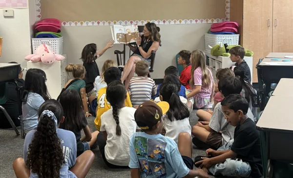 A teacher sits in a chair reading a picture book aloud to a group of children seated on the floor of an elementary school classroom.