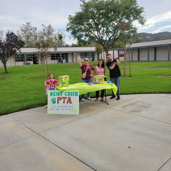 Three adults and a child wave at the camera while standing behind a green table with a Reidy Creek PTA sign and snacks on a school campus.