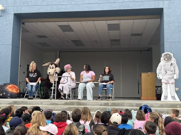 Four adults and a person in a dog mascot costume sit on an outdoor school stage in front of a large audience of children, with a cardboard astronaut cutout and a podium visible nearby.