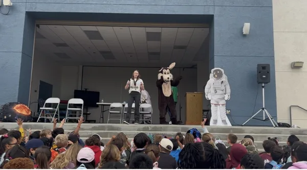 A woman stands on an outdoor school stage with a dog mascot costume character and an astronaut cardboard cutout while a large crowd of elementary school children sit on the ground in front of them with hands raised.