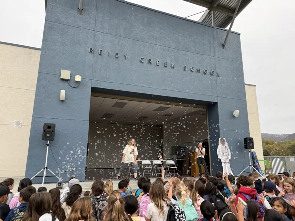 Two presenters perform a bubble show on the outdoor stage of Reidy Creek School for a large crowd of seated elementary school children.