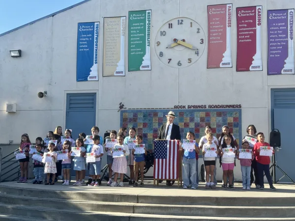 A group of elementary school students hold certificates while standing outside Rock Springs Roadrunners school in front of Character Counts banners, alongside an adult in a suit and hat standing near an American flag.