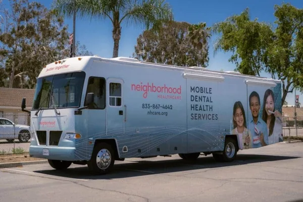 A large mobile dental health services vehicle for Neighborhood Healthcare, with "better together," "neighborhood HEALTHCARE," "833-867-4642," "nhcare.org," and "MOBILE DENTAL HEALTH SERVICES" written on its exterior, parked in a sunny lot with palm trees in the background.